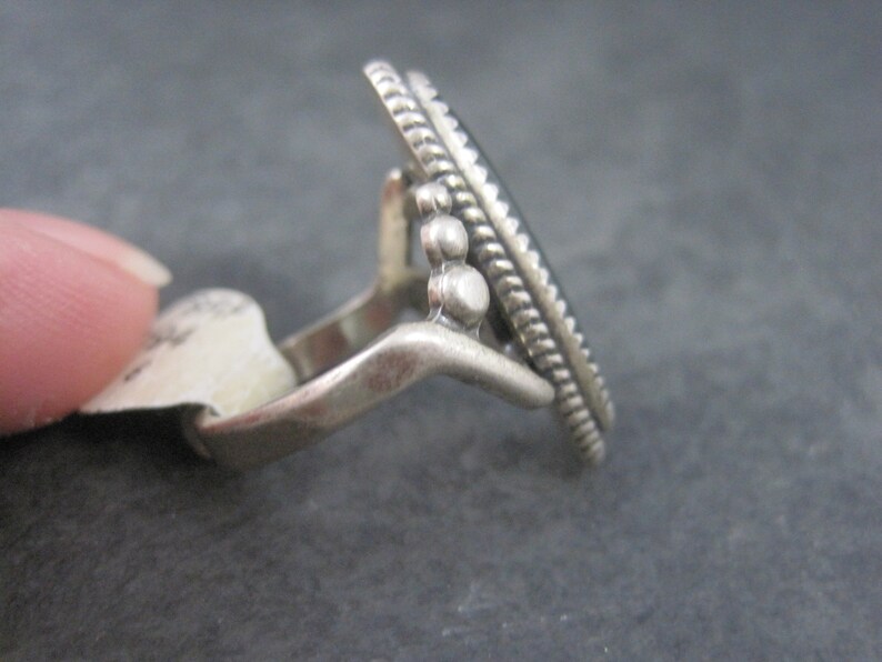 May include: A silver ring with an oval, dark-colored stone. The ring has a decorative, textured border and small, round accents. The ring is shown from a side angle, highlighting its design and craftsmanship. The ring is a piece of jewelry.