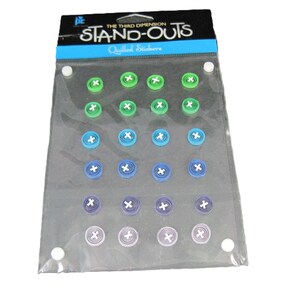 May include: A package of "STAND-OUTS" Quilled Stickers in a variety of colors, including green, blue, and purple. Each sticker has a spiral design with a white cross in the center. The package is sealed in clear plastic.