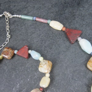 May include: A multi-colored gemstone necklace with a silver clasp. The necklace features various shapes and sizes of stones, including red, brown, white, and blue.