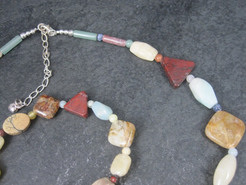 May include: A multi-colored gemstone necklace with a silver clasp. The necklace features various shapes and sizes of stones, including red, brown, white, and blue.