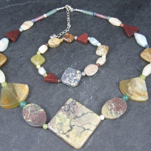 May include: A multi-colored gemstone necklace and bracelet set. The necklace features a large, square-shaped pendant with brown, green, and white marbling. The bracelet has smaller, round and oval-shaped gemstones in various colors, including red, brown, green, and white.