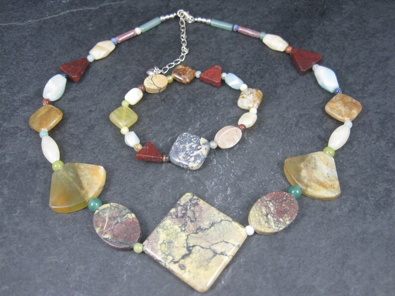 May include: A multi-colored gemstone necklace and bracelet set. The necklace features a large, square-shaped pendant with brown, green, and white marbling. The bracelet has smaller, round and oval-shaped gemstones in various colors, including red, brown, green, and white.
