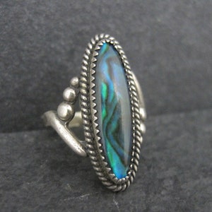 May include: A silver ring with an oval abalone shell inlay. The shell displays iridescent blues, greens, and purples. The ring has a twisted rope border and decorative silver accents.