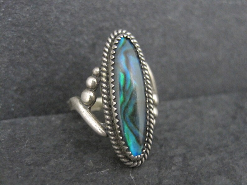 May include: A silver ring with an oval abalone shell inlay. The shell displays iridescent blues, greens, and purples. The ring has a twisted rope border and decorative silver accents.