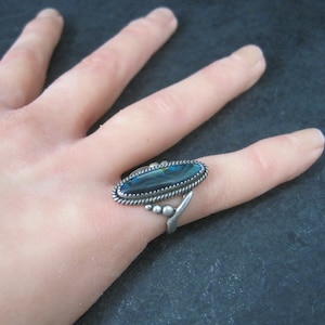 May include: Silver ring featuring an oval, iridescent abalone shell. The ring has a twisted rope border and a decorative design on the band. The abalone shell displays shades of blue, green, and black.