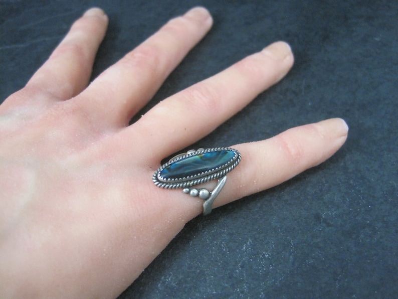 May include: Silver ring featuring an oval, iridescent abalone shell. The ring has a twisted rope border and a decorative design on the band. The abalone shell displays shades of blue, green, and black.