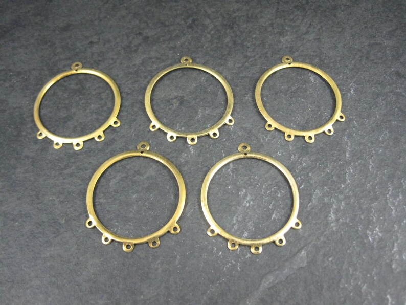 Puede incluir: Cinco componentes de pendientes de metal color lat&oacute;n. Cada componente es un anillo circular con un peque&ntilde;o lazo en la parte superior y seis lazos m&aacute;s peque&ntilde;os espaciados uniformemente en el borde inferior. Los componentes est&aacute;n dispuestos sobre una superficie gris oscuro.