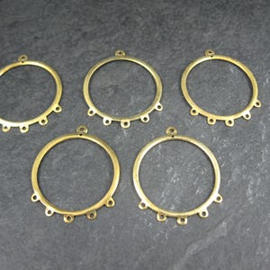 Puede incluir: Cinco componentes de pendientes de metal color lat&oacute;n. Cada componente es un anillo circular con un peque&ntilde;o lazo en la parte superior y seis lazos m&aacute;s peque&ntilde;os espaciados uniformemente en el borde inferior. Los componentes est&aacute;n dispuestos sobre una superficie gris oscuro.