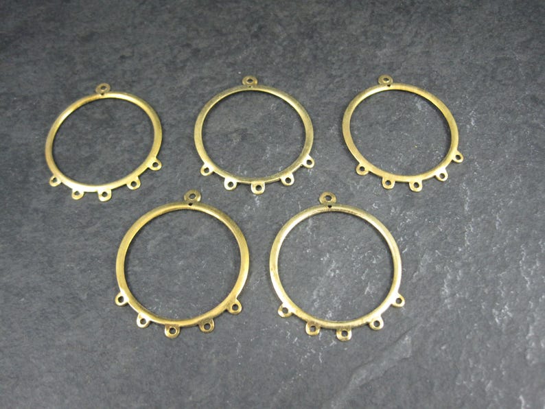 Puede incluir: Cinco componentes de pendientes de metal color lat&oacute;n. Cada componente es un anillo circular con un peque&ntilde;o lazo en la parte superior y seis lazos m&aacute;s peque&ntilde;os espaciados uniformemente en el borde inferior. Los componentes est&aacute;n dispuestos sobre una superficie gris oscuro.