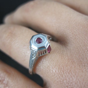 May include: A silver ring with a red gemstone in the center and smaller red gemstones on the sides. The ring is set with small white gemstones around the center stone.