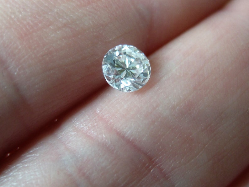 May include: A clear round gemstone with facets, set against a light pink background.
