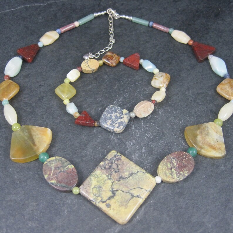 May include: A multi-colored gemstone necklace and bracelet set. The necklace features a large, square-shaped pendant with a marbled pattern in shades of brown, yellow, and green. The bracelet is made of smaller, round and square-shaped gemstones in various colors, including red, brown, yellow, green, and blue.