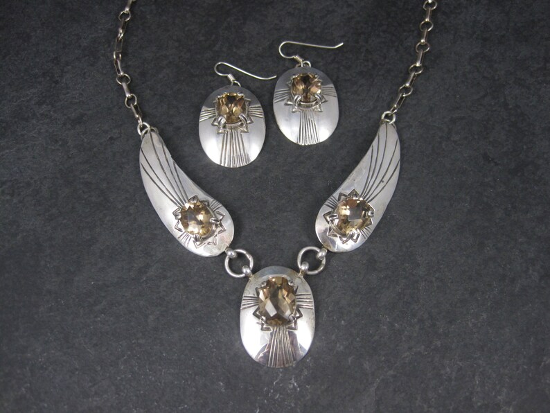 May include: A silver necklace and earrings set with three oval-shaped topaz gemstones. The necklace features a delicate chain and two curved pendants with intricate designs. The earrings are simple and elegant, with a single topaz gemstone hanging from each.