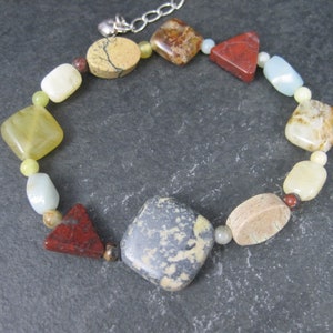 May include: A multi-colored gemstone bracelet with a silver clasp. The bracelet features a variety of shapes and sizes of stones, including square, rectangular, and triangular stones. The stones are in shades of brown, red, yellow, and white.