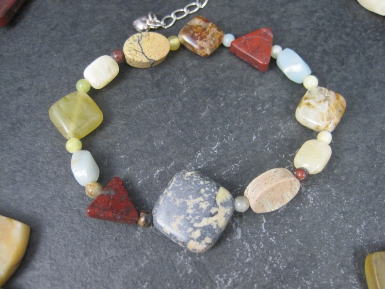 May include: A multi-colored gemstone bracelet with a silver clasp. The bracelet features a variety of shapes and sizes of stones, including square, rectangular, and triangular stones. The stones are in shades of brown, red, yellow, and white.