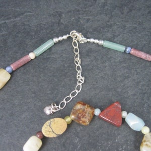 May include: A multi-colored gemstone necklace with a silver clasp and extender chain. The necklace features various shapes and sizes of stones, including red, brown, green, blue, and white.