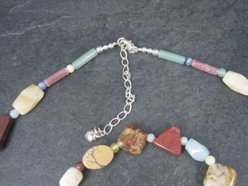 May include: A multi-colored gemstone necklace with a silver clasp and extender chain. The necklace features various shapes and sizes of stones, including red, brown, green, blue, and white.