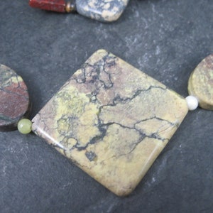 May include: A close-up of a square-shaped, light brown, yellow, and gray gemstone with a marbled pattern. The gemstone is part of a necklace with other colorful gemstones.