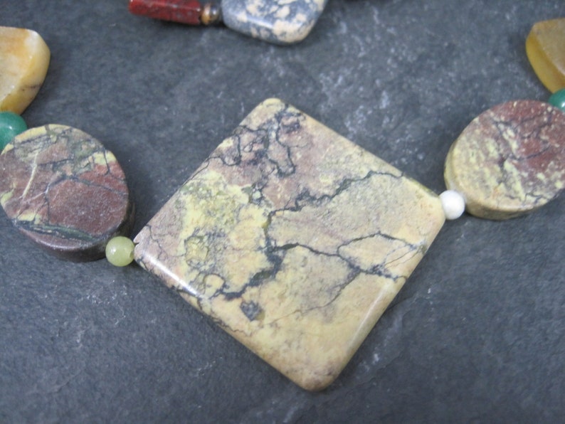 May include: A close-up of a square-shaped, light brown, yellow, and gray gemstone with a marbled pattern. The gemstone is part of a necklace with other colorful gemstones.