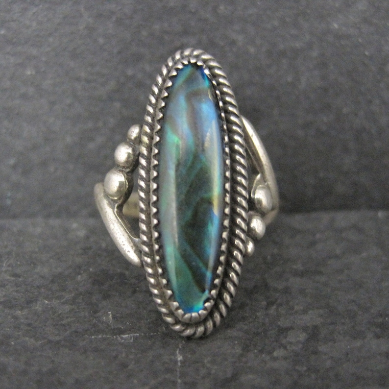 May include: A silver ring with an oval abalone shell center. The shell has iridescent blue, green, and purple hues. The ring has a twisted rope border and decorative silver accents on the sides.