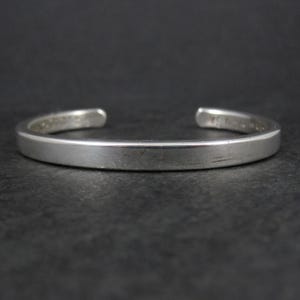 May include: A silver cuff bracelet with a simple, minimalist design. The open-ended bracelet is made of a solid, polished metal, and is displayed against a dark background. This jewelry item is suitable for everyday wear.