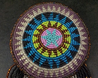 May include: A beaded Native American dreamcatcher with a circular design featuring a central flower motif in shades of pink, blue, and green. The outer ring is a geometric pattern in shades of brown, purple, and white. The dreamcatcher has black and silver fringe.