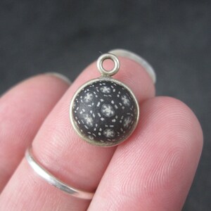 May include: A round pendant with a dark background and a pattern of white floral shapes. The pendant has a silver-colored metal frame and a loop at the top for attaching to a chain or bracelet. The pendant is held between fingers.