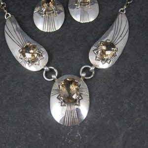 May include: A silver necklace with a teardrop-shaped pendant and matching earrings. The pendant and earrings feature a large, faceted, topaz-colored gemstone set in a silver starburst design.