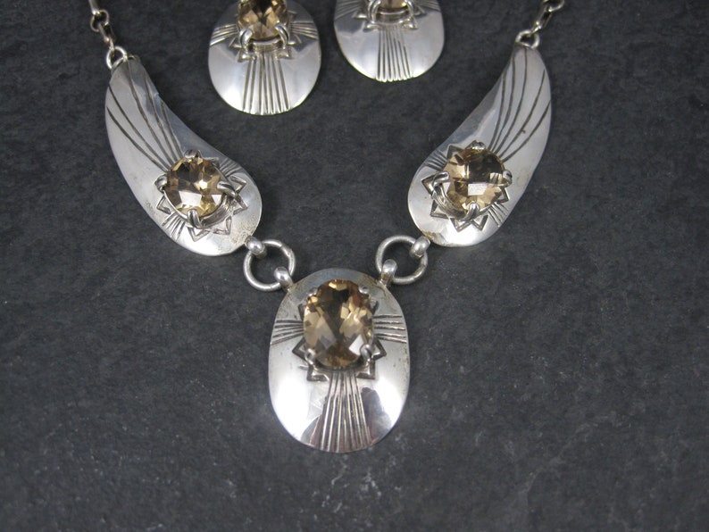 May include: A silver necklace with a teardrop-shaped pendant and matching earrings. The pendant and earrings feature a large, faceted, topaz-colored gemstone set in a silver starburst design.