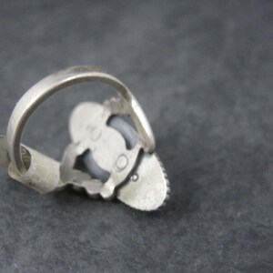 May include: A silver-toned ring with a decorative setting. The ring features a central, oval-shaped stone, surrounded by smaller stones. The band is simple and smooth. The ring is shown from an angle, highlighting its design and craftsmanship.