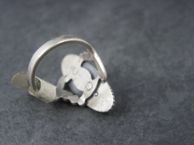 May include: A silver-toned ring with a decorative setting. The ring features a central, oval-shaped stone, surrounded by smaller stones. The band is simple and smooth. The ring is shown from an angle, highlighting its design and craftsmanship.