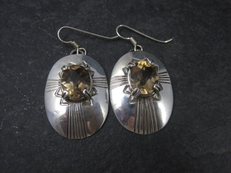 May include: A pair of silver earrings with oval-shaped settings featuring a brown gemstone. The earrings have a geometric design with a starburst pattern.