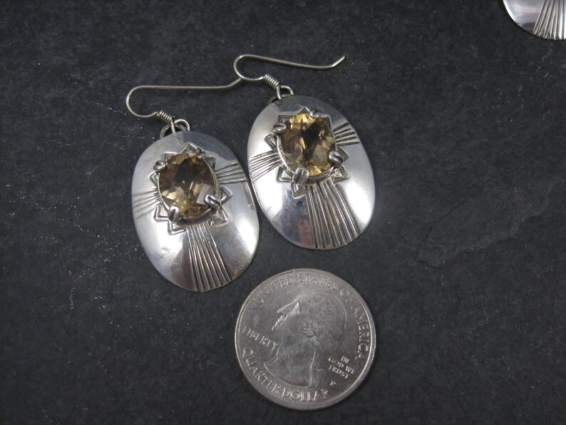 May include: A pair of silver earrings with oval-shaped settings featuring a brown gemstone. The earrings have a geometric design with radiating lines.