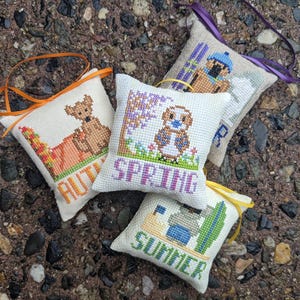 Op de afbeelding: Vier kleine, vierkante, witte stoffen kussentjes met geborduurde afbeeldingen en tekst die de vier seizoenen voorstellen. De kussentjes zijn versierd met oranje, gele, groene, blauwe en paarse draad. De kussentjes zijn in een halve cirkel op een bruin en grijs grind oppervlak gerangschikt. De tekst op de kussentjes luidt "Lente", "Zomer", "Herfst" en "Winter".