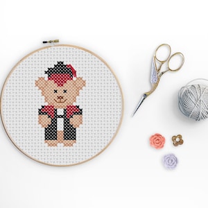 Könnte beinhalten: Ein Kreuzstich-Stickrahmen mit einem Motiv eines Bären, der eine Kappe und eine Robe trägt. Der Rahmen befindet sich auf einer weißen Oberfläche mit einer Garnrolle, einer Schere und dekorativen Blumen.