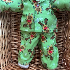 Peut inclure: Une poupée portant une chemise à manches longues boutonnée vert clair et un pantalon assorti avec un motif répétitif de rennes marron au nez rouge emmêlés dans des guirlandes lumineuses et des flocons de neige blancs. Cheveux brun foncé.