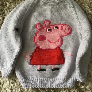 May include: A pale blue knitted jumper featuring a pink pig character in a red dress. The pig is smiling and is the central design. The jumper has long sleeves and a ribbed neckline. The garment is hand-knitted.