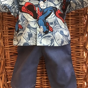 May include: A doll wearing a blue button-down shirt with a Spider-Man comic book print and blue pants. The shirt has a collar and is buttoned up. The pants are a solid blue color.