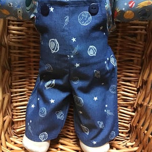 May include: A pair of dark blue denim dungarees featuring a white line-art pattern of planets, stars, and rockets, paired with a collared shirt underneath that has a colourful space-themed print of planets, rockets, and stars on a light blue background.