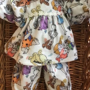 May include: A doll wearing a white pajama set with a colorful cartoon print. The pajamas feature a top with a lace collar and matching pants. The cartoon characters are in various poses and colors.