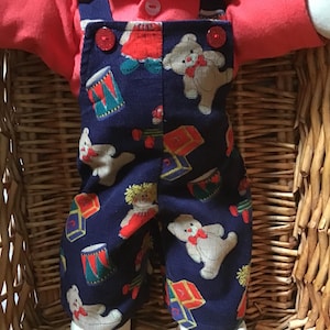 May include: A doll wearing a red collared shirt and navy blue dungarees. The dungarees feature a repeating pattern of teddy bears, toy drums, and alphabet blocks. The dungarees have two red buttons at the top of the bib and a white button on the shirt.