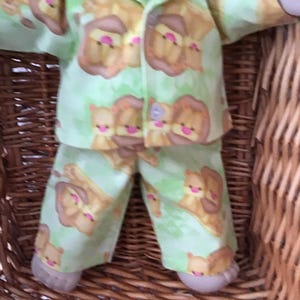 May include: Green pajamas with a brown lion print for a doll. The pajamas have a button-up front and long sleeves.