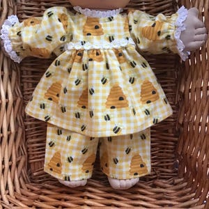 May include: A doll dressed in a yellow and white gingham outfit featuring a bee and beehive pattern. The outfit comprises a top with a frilled collar and sleeves, and matching trousers. The doll is placed in a wicker basket.