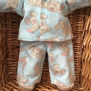 May include: A light blue pajama set featuring a pattern of tan bunnies with pink ears. The top has a button-down front and collar. The straight-legged trousers and top are on a doll. The pajamas are made of soft fabric.