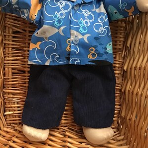 May include: A blue and white shirt with a shark and wave pattern. The shirt has a button-up front and long sleeves. The doll is wearing navy blue corduroy pants.