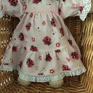 May include: A pink doll's dress with a ladybug print and white lace trim. The dress has a ruffled skirt and a pink ribbon around the waist.