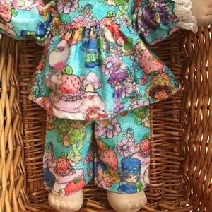 May include: A doll with dark brown yarn hair wearing a light blue long-sleeved top and matching pants featuring a colorful print of strawberries, flowers, and small characters. The top has a white lace collar and white lace cuffs on the sleeves.
