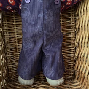May include: A purple corduroy overall with a subtle jack-o'-lantern print, featuring two matching purple buttons at the chest. Underneath, a collared shirt displays a vibrant pattern of orange pumpkins on a dark purple background.