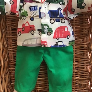 May include: A doll wearing a button-down shirt and green corduroy pants. The shirt is white with a colorful pattern of various vehicles, including trucks and cars. The green pants are a solid color. The doll is in a wicker basket.
