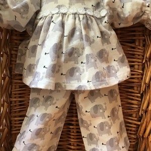 May include: A doll wearing a grey and white pajama set with an elephant print. The pajamas have a white lace trim and a white flower trim around the neckline.